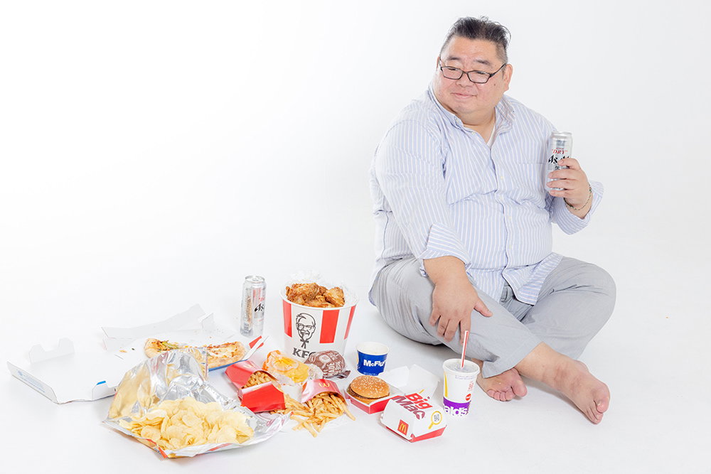 揚げ物やハンバーガーなどに囲まれてほっこりする肥満男性