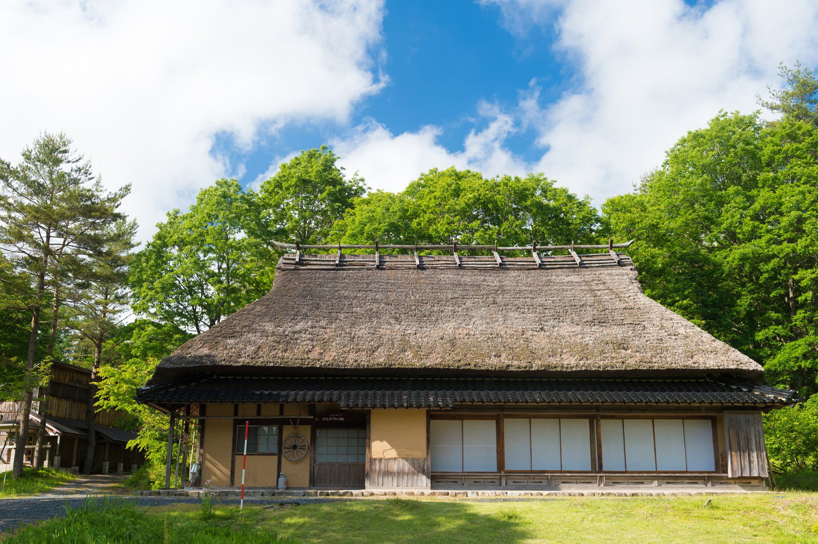 青空と緑の木々に囲まれた茅葺屋根の古民家