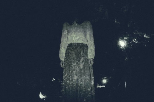 仄暗い闇の奥から立つ白い衣服の女性の怪奇的な人影
