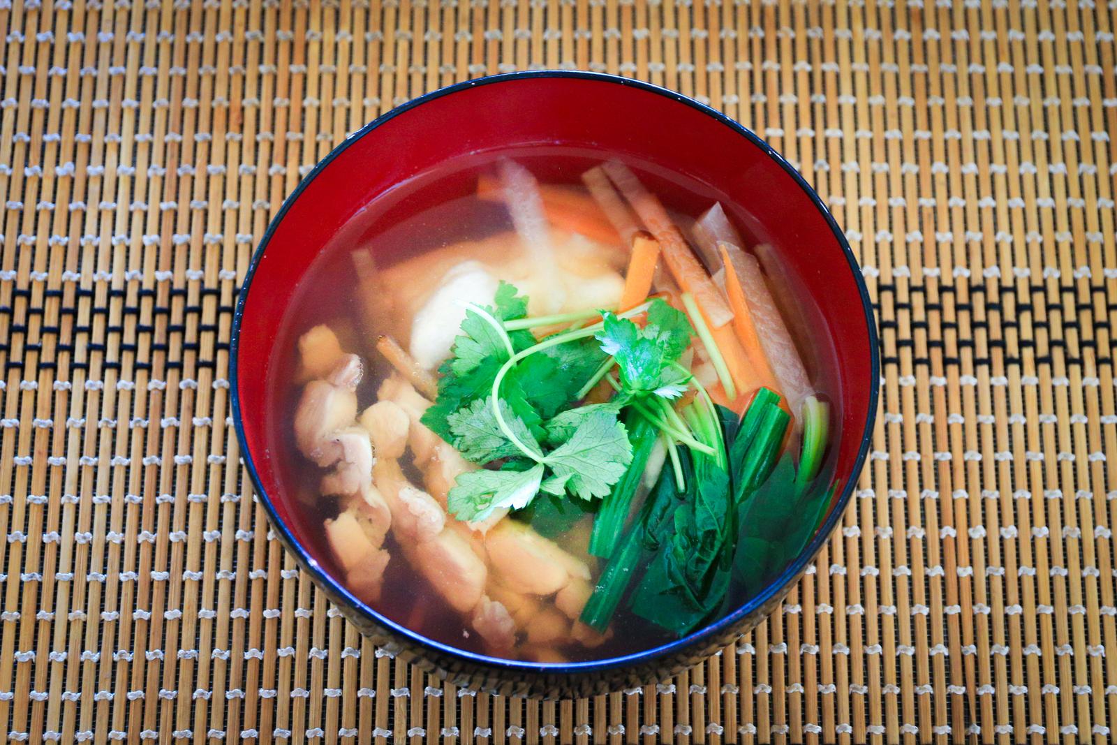 Clear broth zoni with mitsuba and chicken served in a red lacquered bowl placed on a bamboo mat