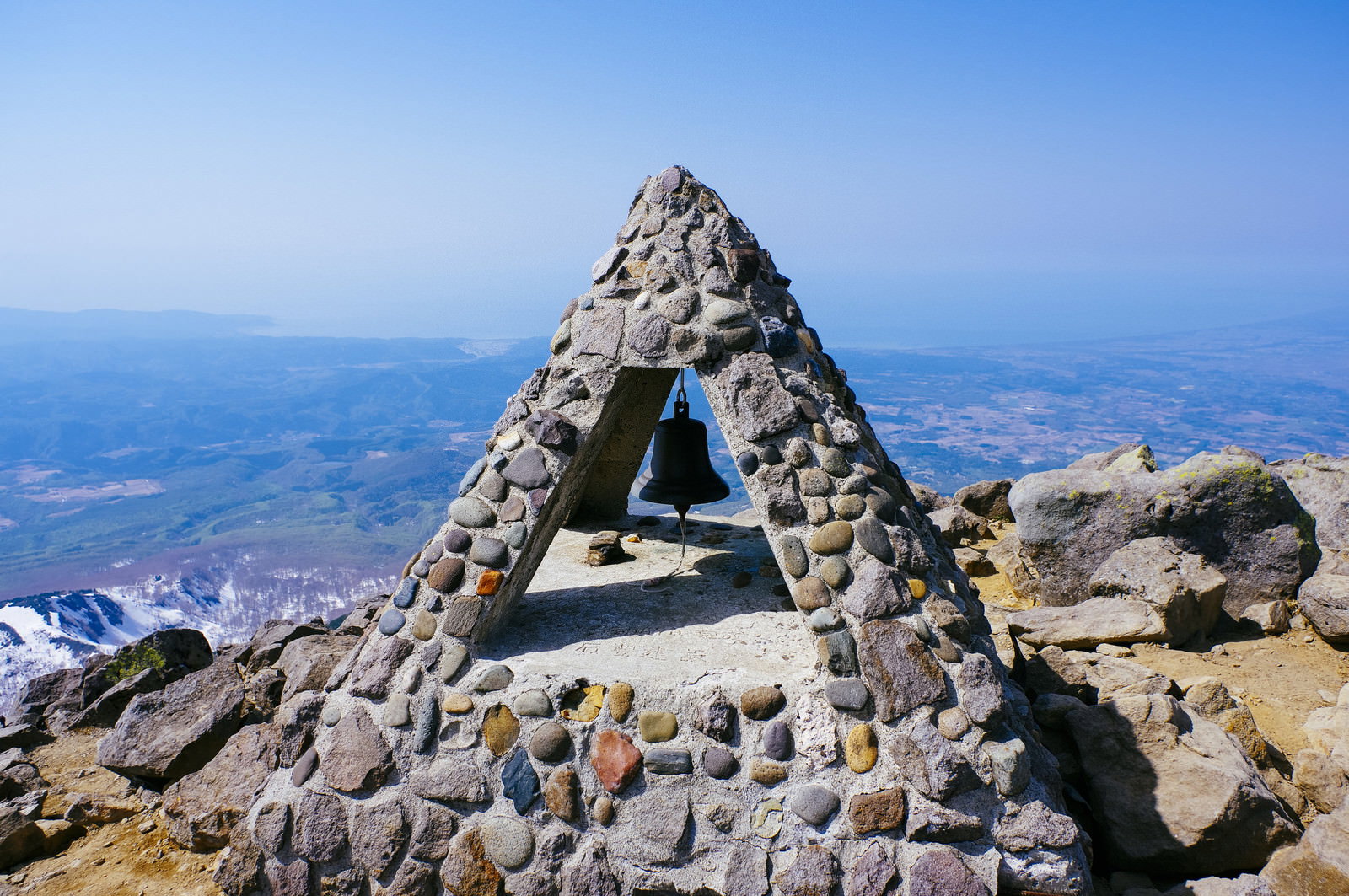 青空の下、岩木山山頂に建つ石積みの三角形の碑と中央に吊り下げられた鐘
