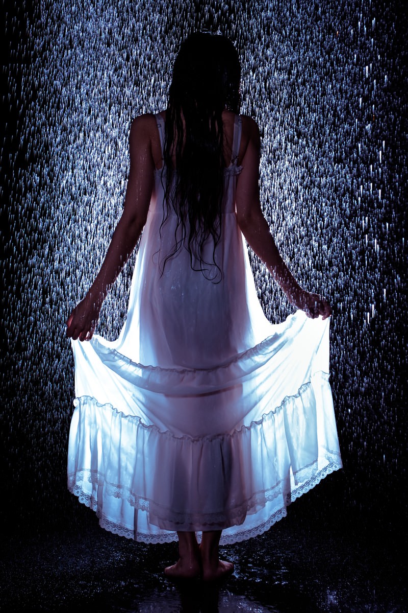 激しい雨の中で白いワンピースの裾をあげる女性の後ろ姿［モデル：緋真煉］