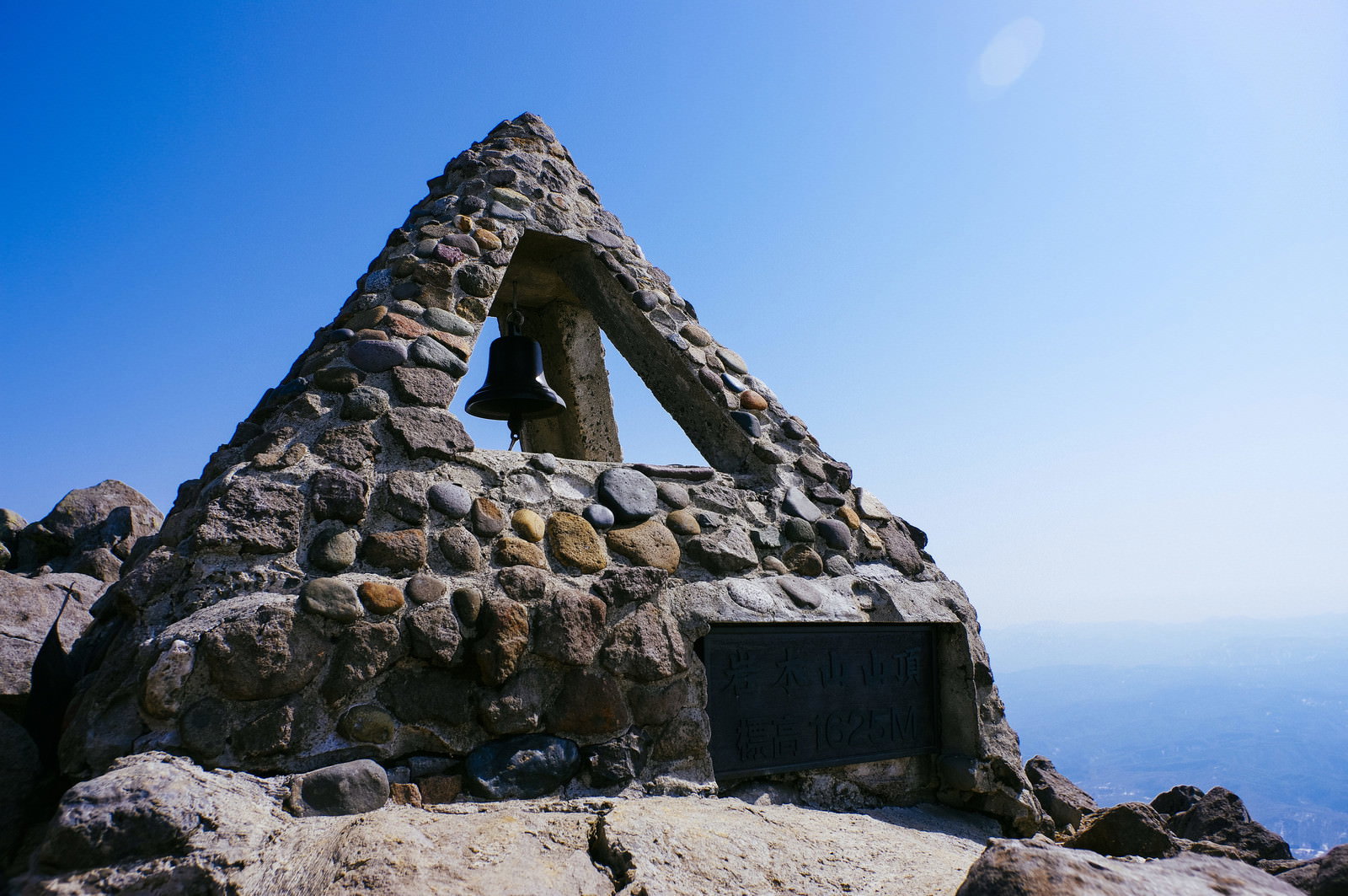 青空の下、岩木山山頂に立つ石組みの三角形の碑と鐘