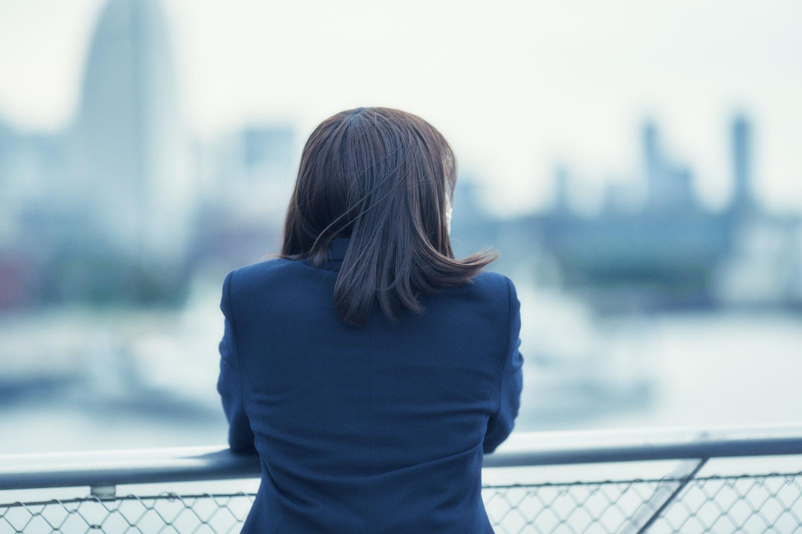 紺色の制服姿で、ビル立ち並ぶ街並みを眺める女子高生の後ろ姿
