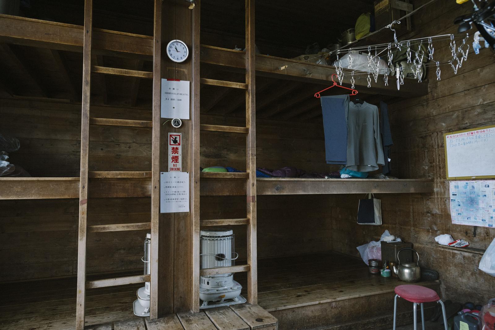 八甲田山の山小屋ヒュッテ内部。木造建物内に衣類ハンガーと生活用具が見える