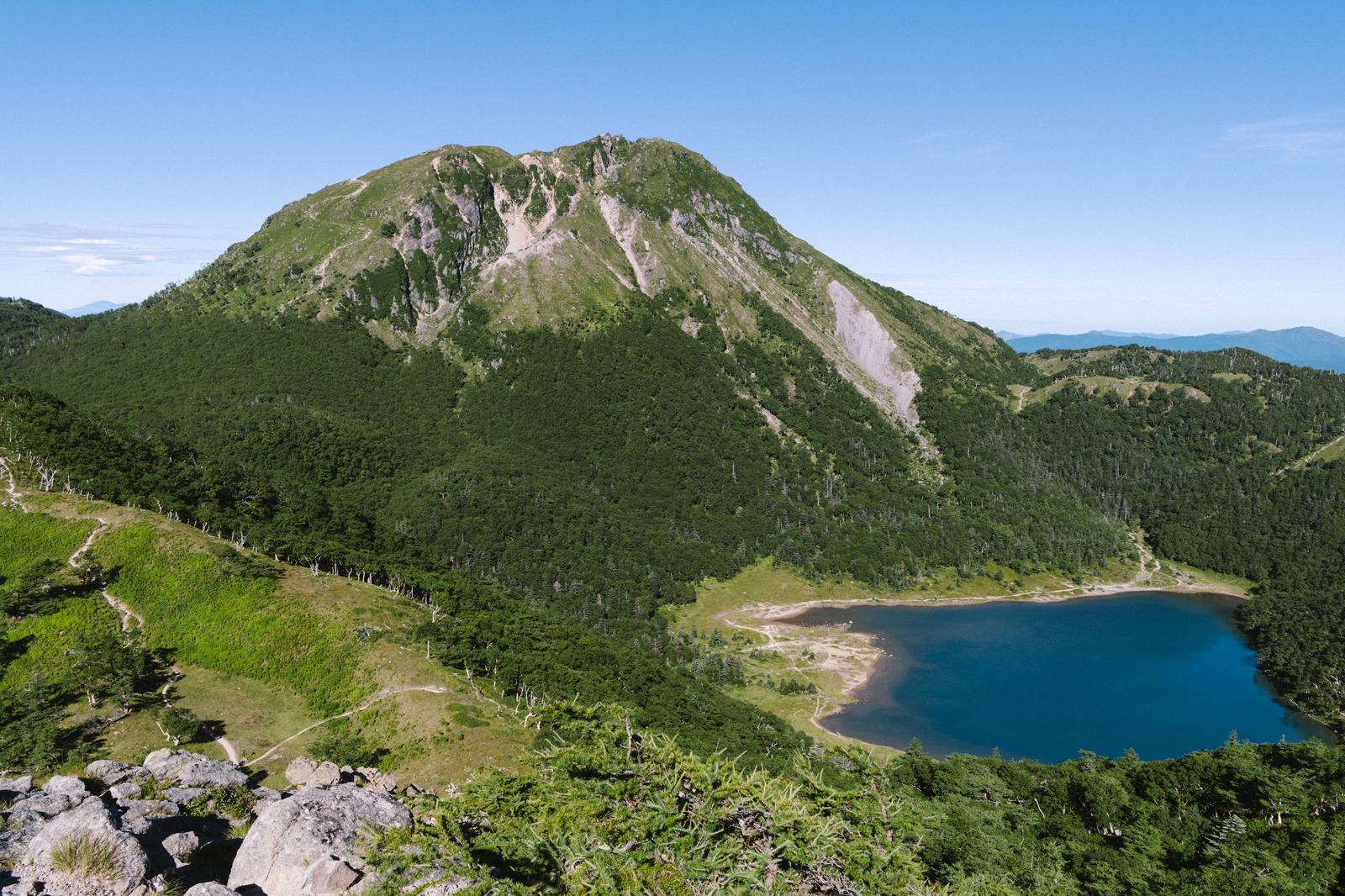 五色沼を望む日光白根山の登山風景。青い湖と緑の森に囲まれた岩山