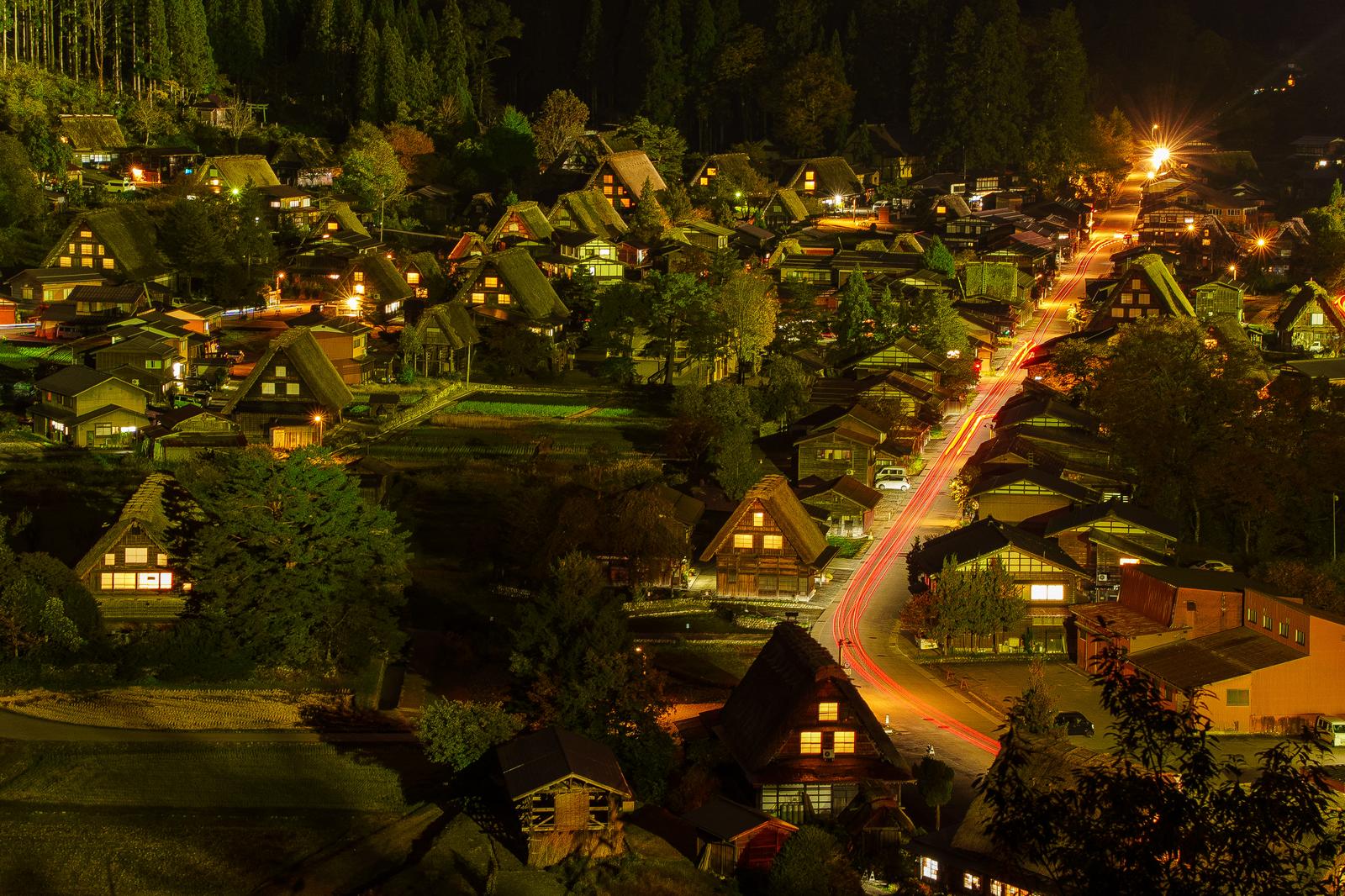 白川郷の合掌造り集落が夜間にライトアップされ、道路に赤い光跡が映る風景