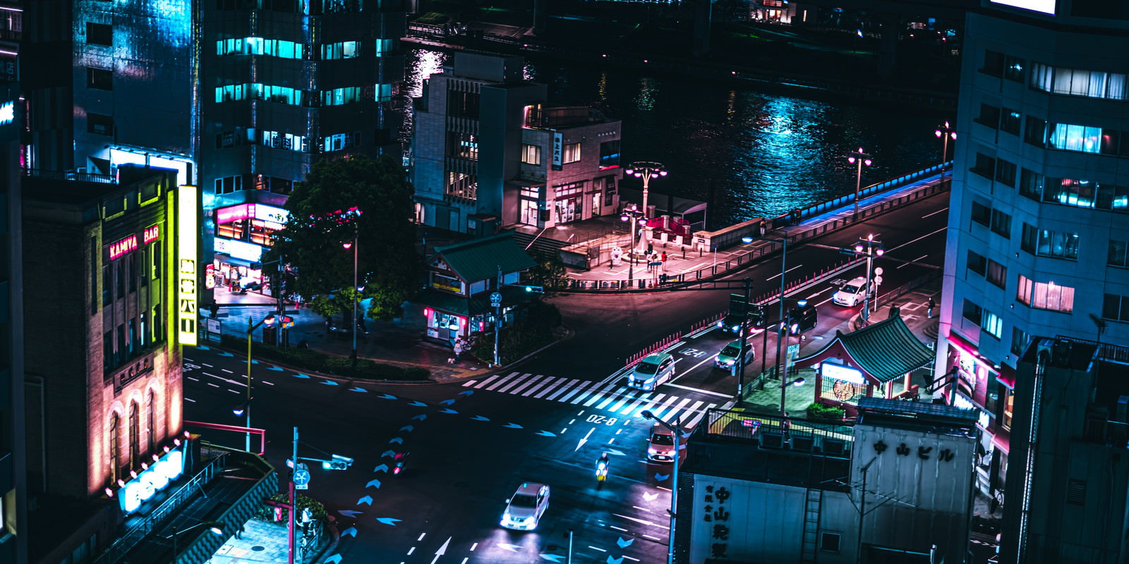 夜の水上バス乗り場前の交差点でネオンサインが光る中を車両が通行する様子