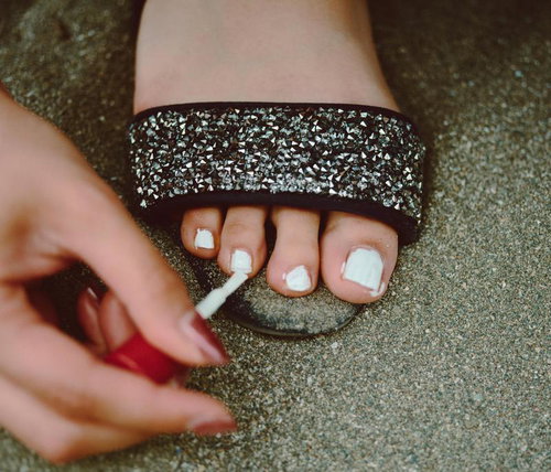 グリッター素材の黒いブレスレットをした足の指に白いマニキュアを塗る