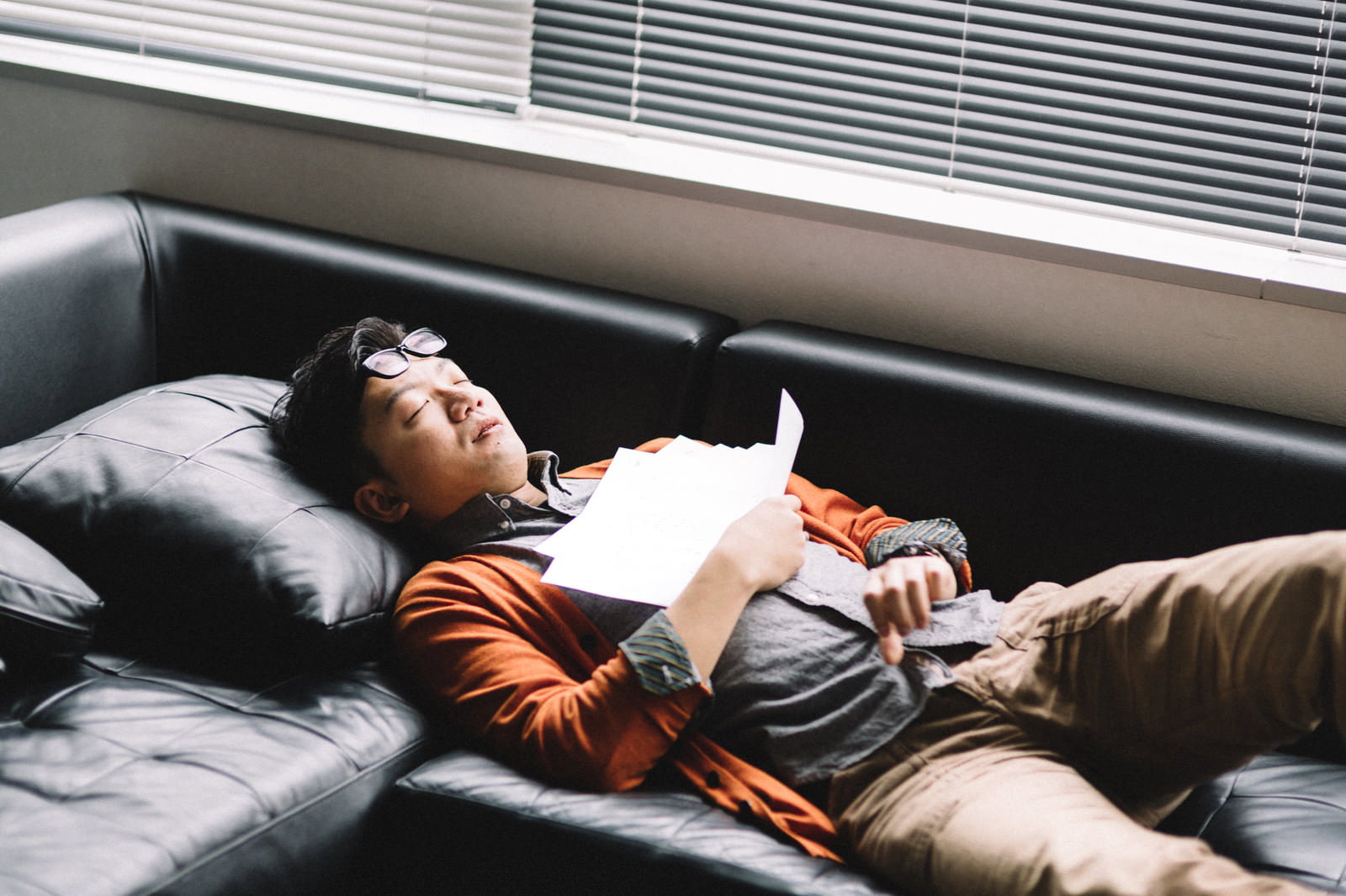 黒いソファーで書類を抱えながら眠るオレンジシャツの男性［モデル：ozpa］