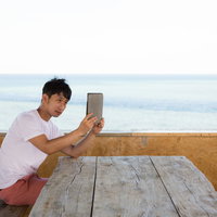 タブレット端末で海を背景に自撮りをする男性の写真