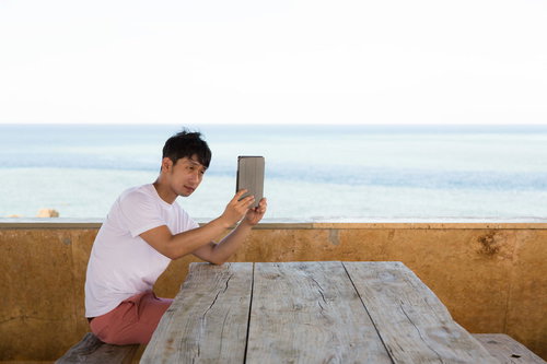 タブレット端末で海を背景に自撮りをする男性