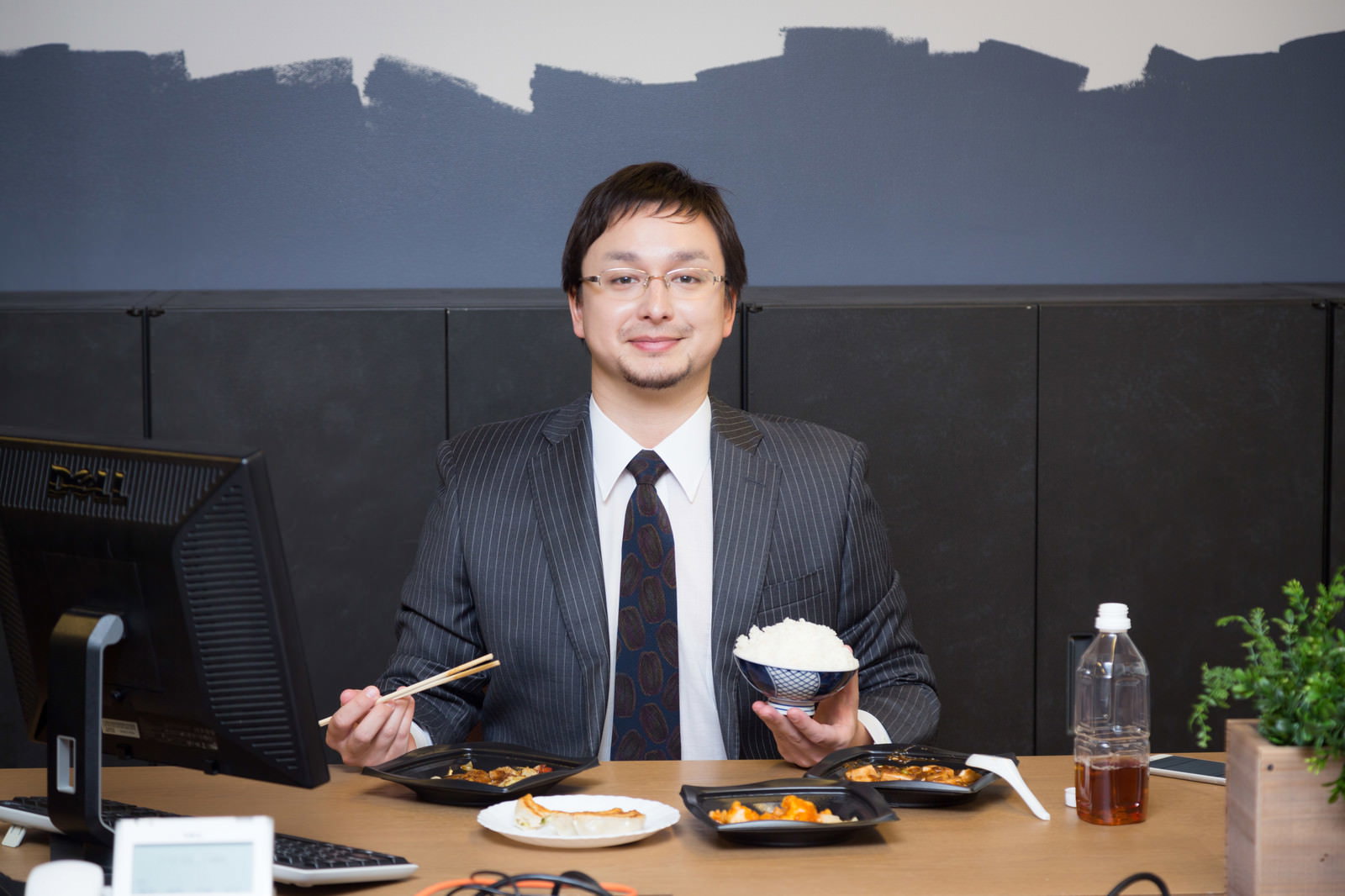 スーツ姿の眼鏡をかけた男性がオフィスのデスクで黒い弁当容器からご飯を箸で食べている［モデル：Max_Ezaki］