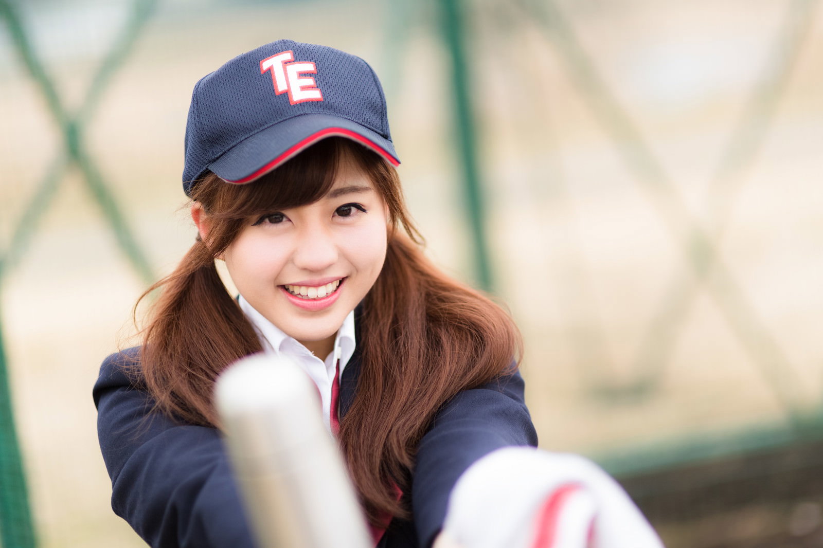 野球部グラウンドで紺色キャップ・紺色ジャージ姿の女子高生がタオルと水筒を持ち笑顔を向ける［モデル：河村友歌］