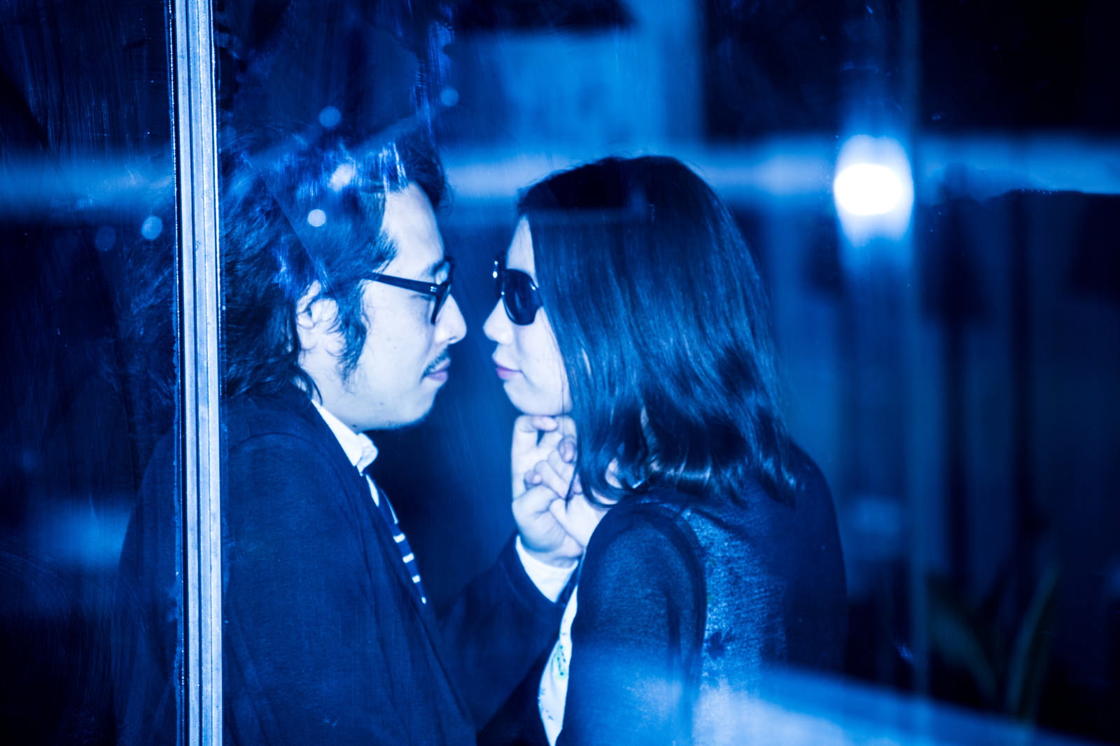 青色照明に包まれた夜の街で、眼鏡の男性とサングラスの女性が向き合う［モデル：ゆうせい］