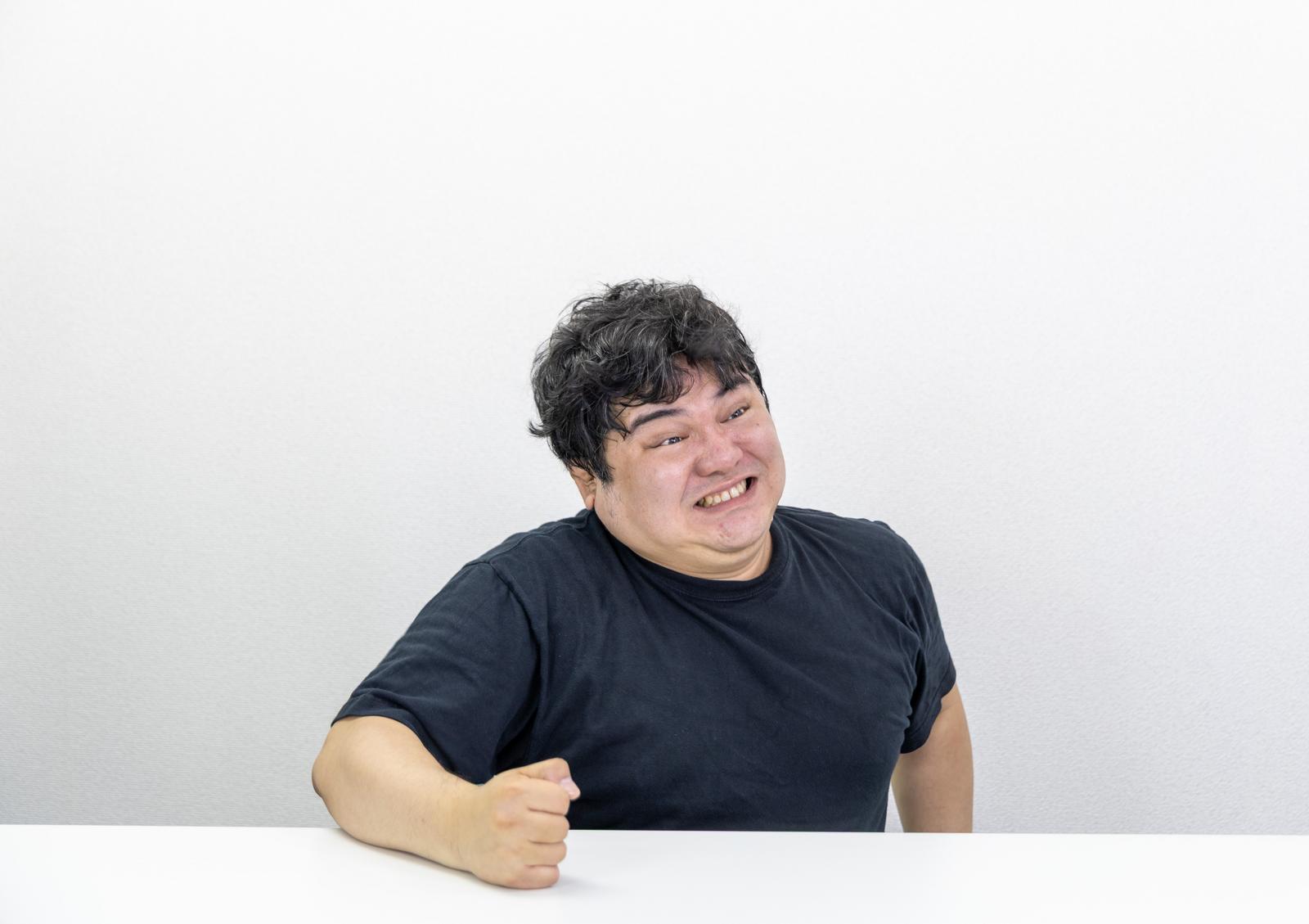黒いTシャツを着た男性が歯を食いしばり握りこぶしで台を叩くポーズ［モデル：段田隼人］