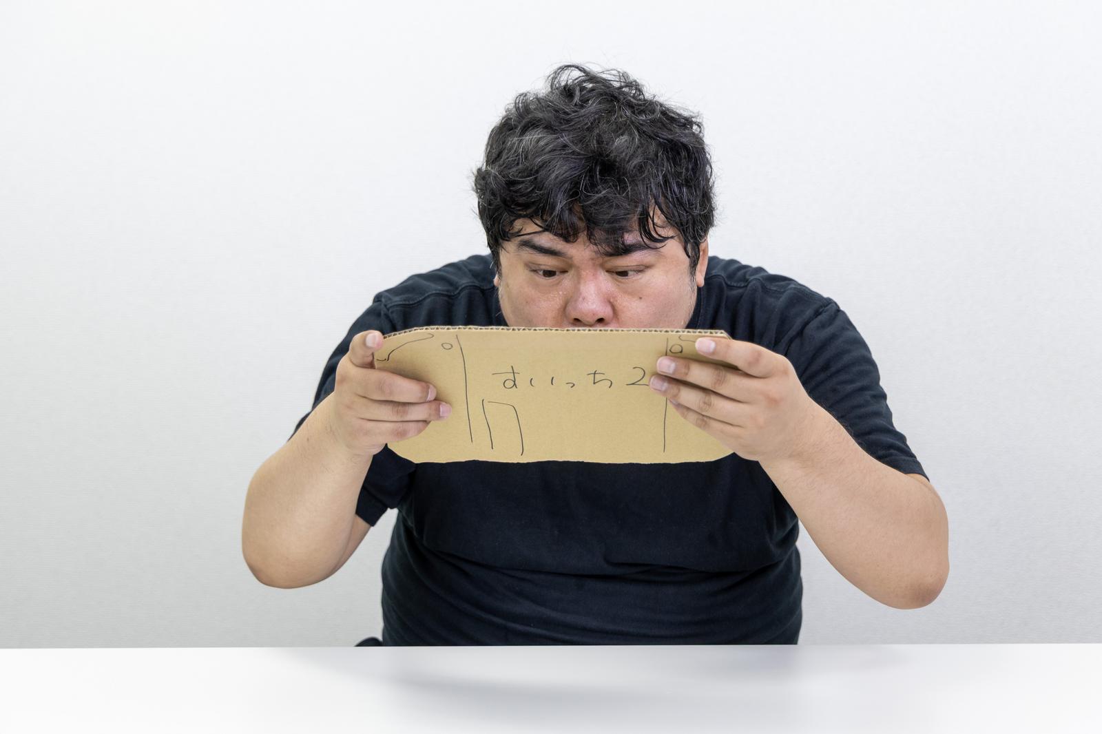 段ボール製のゲーム機を操作し連打の練習をする黒いTシャツの男性［モデル：段田隼人］