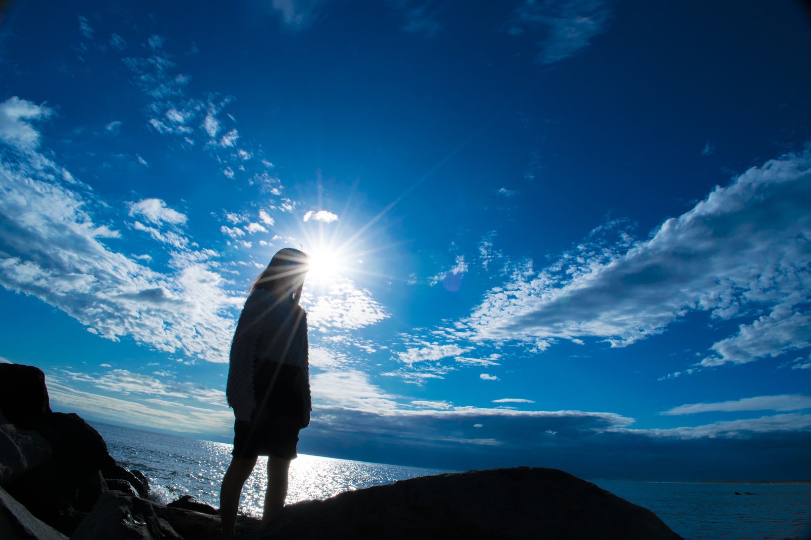 青空と雲が広がる海岸で、強い日差しの中に立つ女性のシルエット