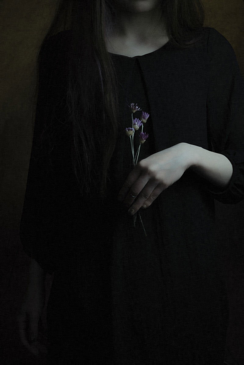 暗い背景で紫色の造花を手に持つ黒ワンピース姿の女性