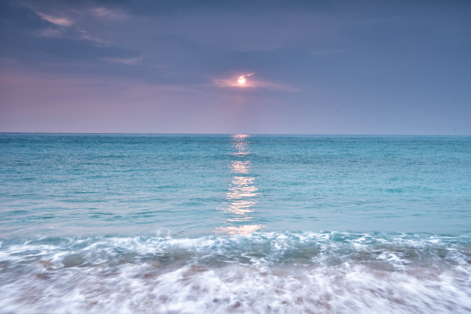 エメラルドグリーンの海に夕日が反射し、白い波が打ち寄せる波打ち際