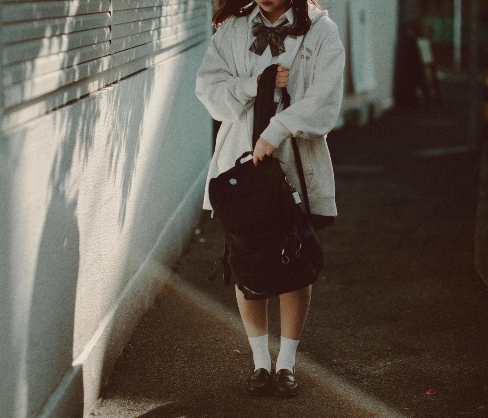 白い壁沿いの通路を歩く、制服姿のリュックを背負った女子高生