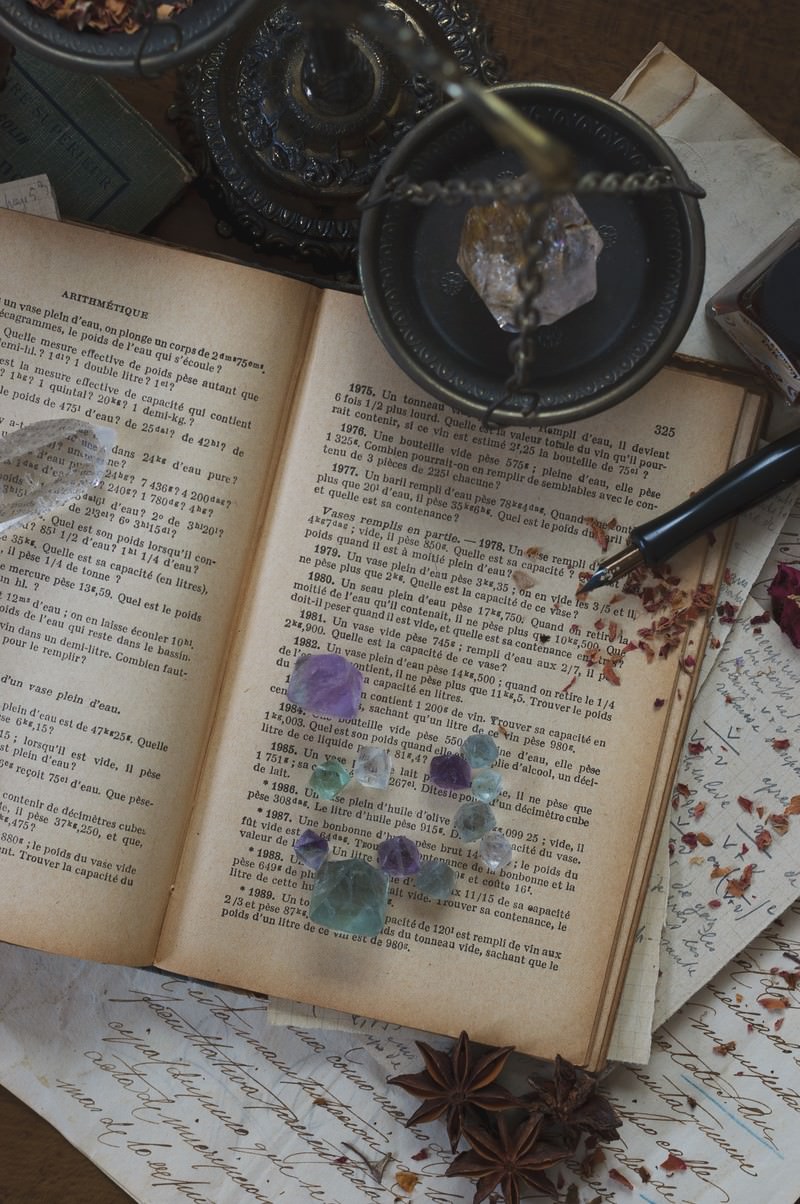 開かれた洋書の上に青い水晶が置かれ、周囲に羽根ペンやルーペがある静物