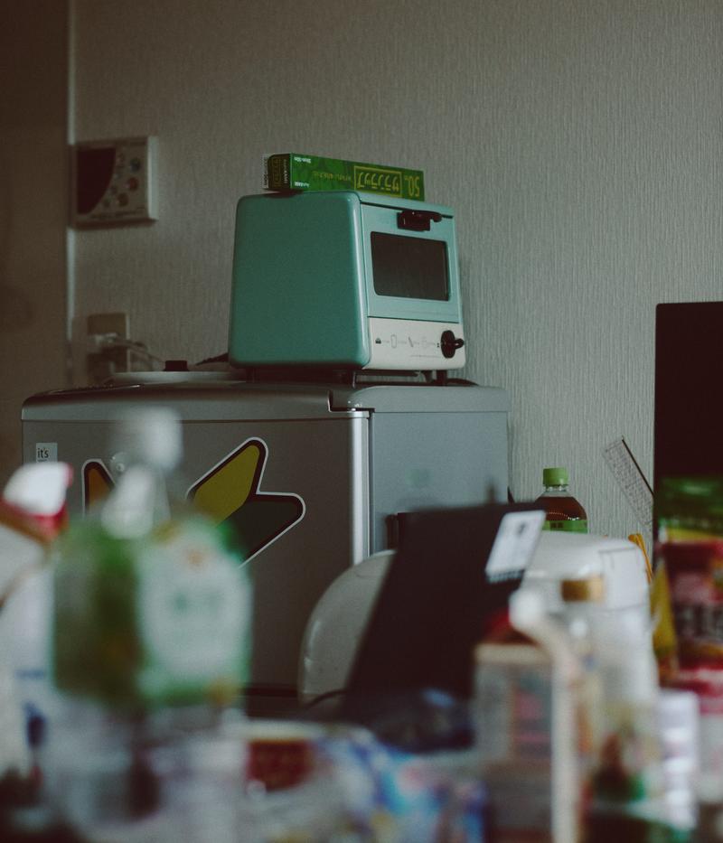 グリーンの小型冷蔵庫の上に白いオーブンレンジが置かれ、手前に食べ物や調味料が散らばるキッチン