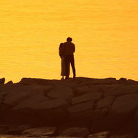 夕焼け空をバックに海岸の岩場でいちゃつくカップルのシルエットの写真