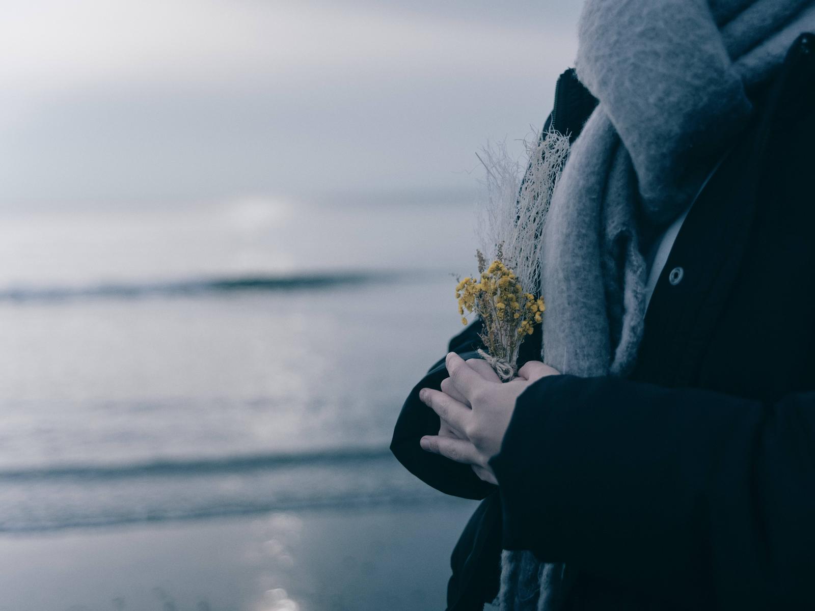 グレーのコートを着た学生がドライフラワーを持ち海辺に立っている