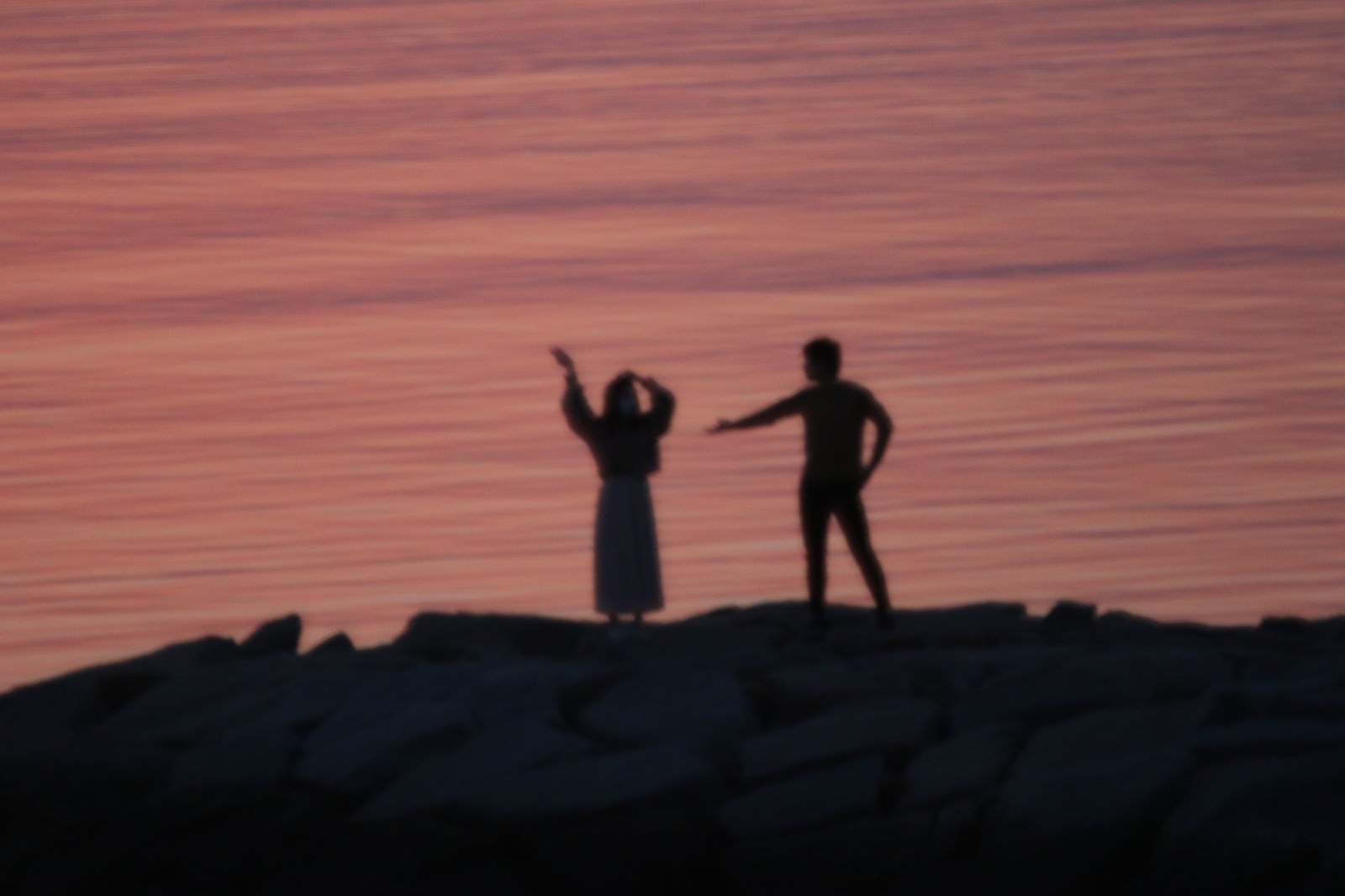 夕暮れの海岸で岩場に立ち両手を上げてはしゃぐカップルのシルエット