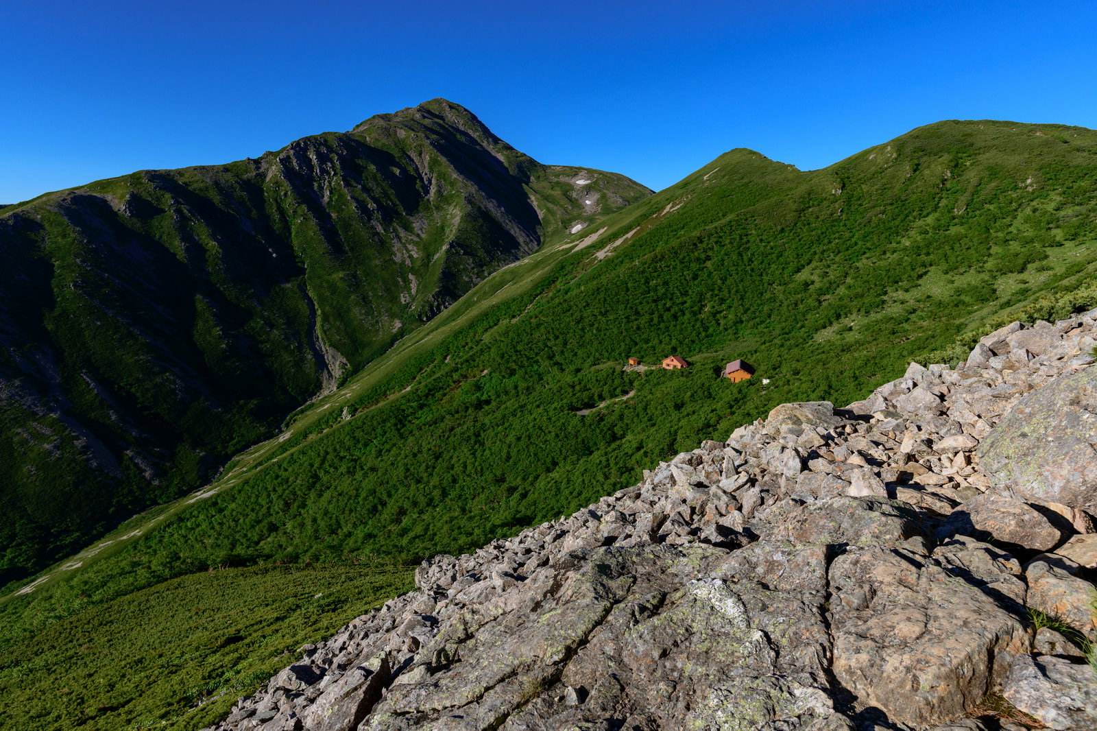 南アルプスの稜線に建つ山小屋と背景の赤石岳、青空が広がる登山風景