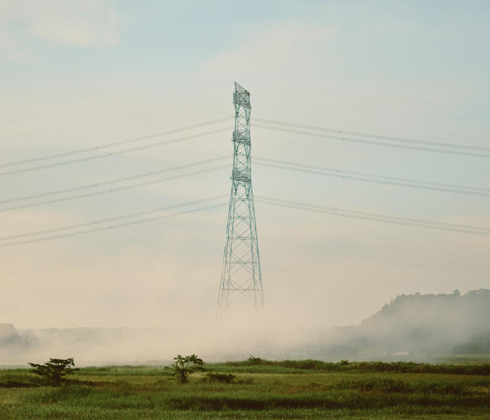 朝霧に覆われた田園地帯に立つ緑色の送電鉄塔と複数の高圧電線