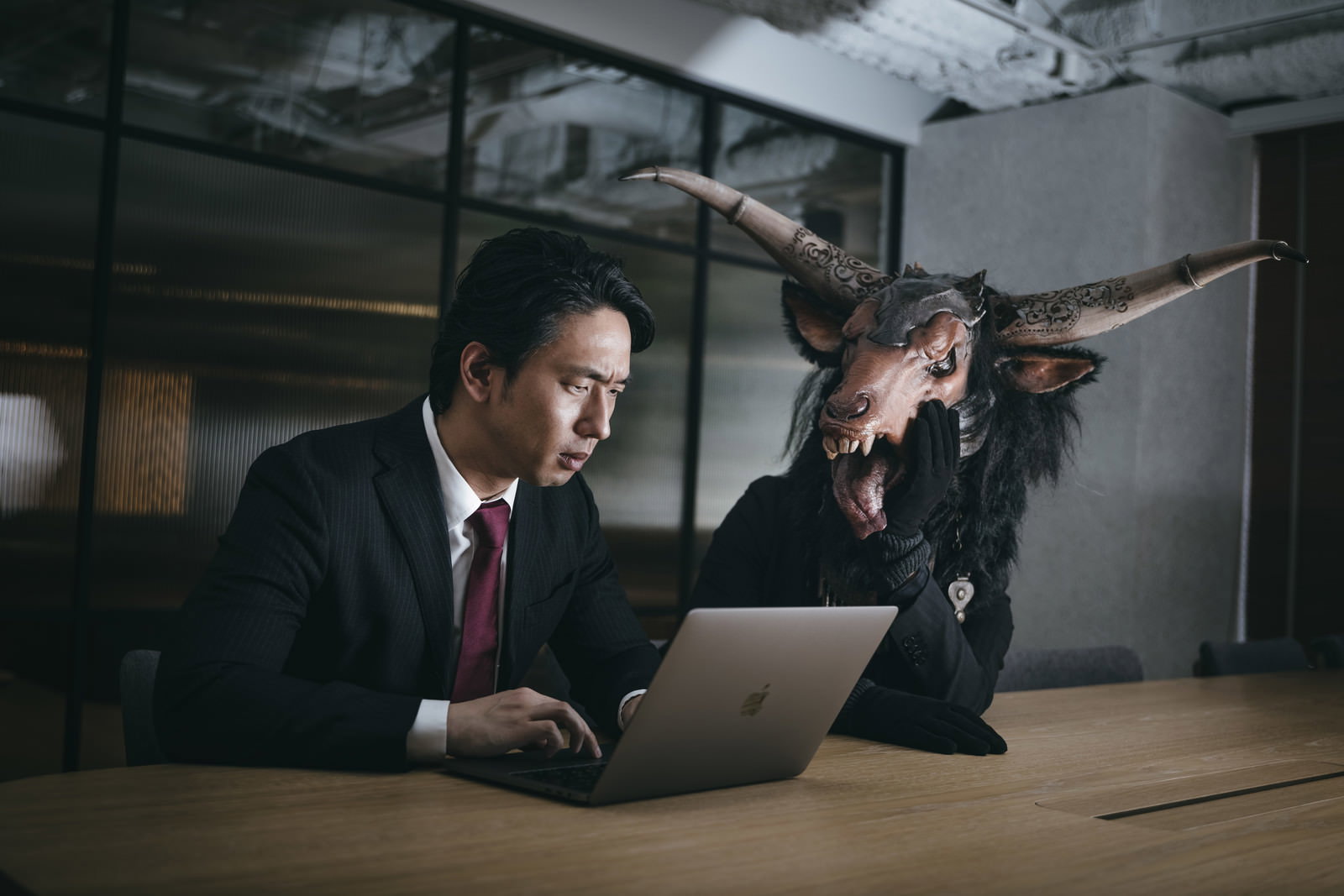 薄暗いオフィスでノートパソコンに向かうスーツ姿の男性と、その隣に立つ悪魔風の存在［モデル：大川竜弥］