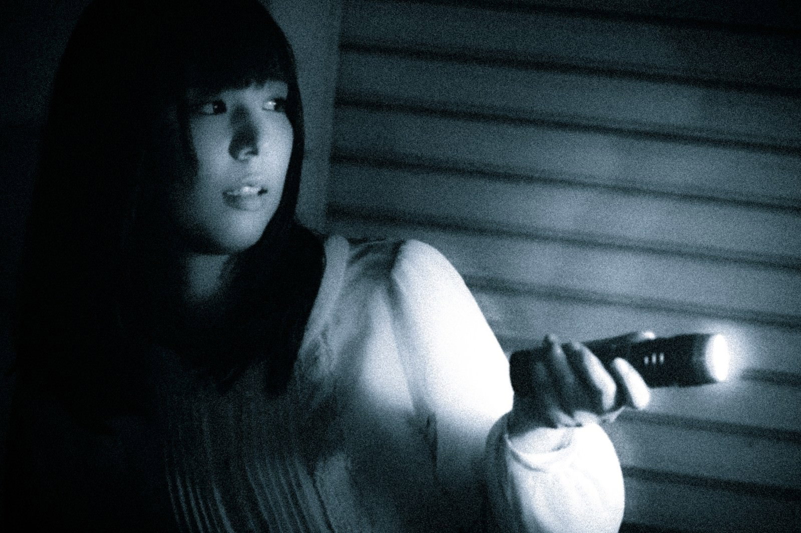 懐中電灯を手にした女性が暗い木製シャッター前で身をすくめている［モデル：さとうゆい］