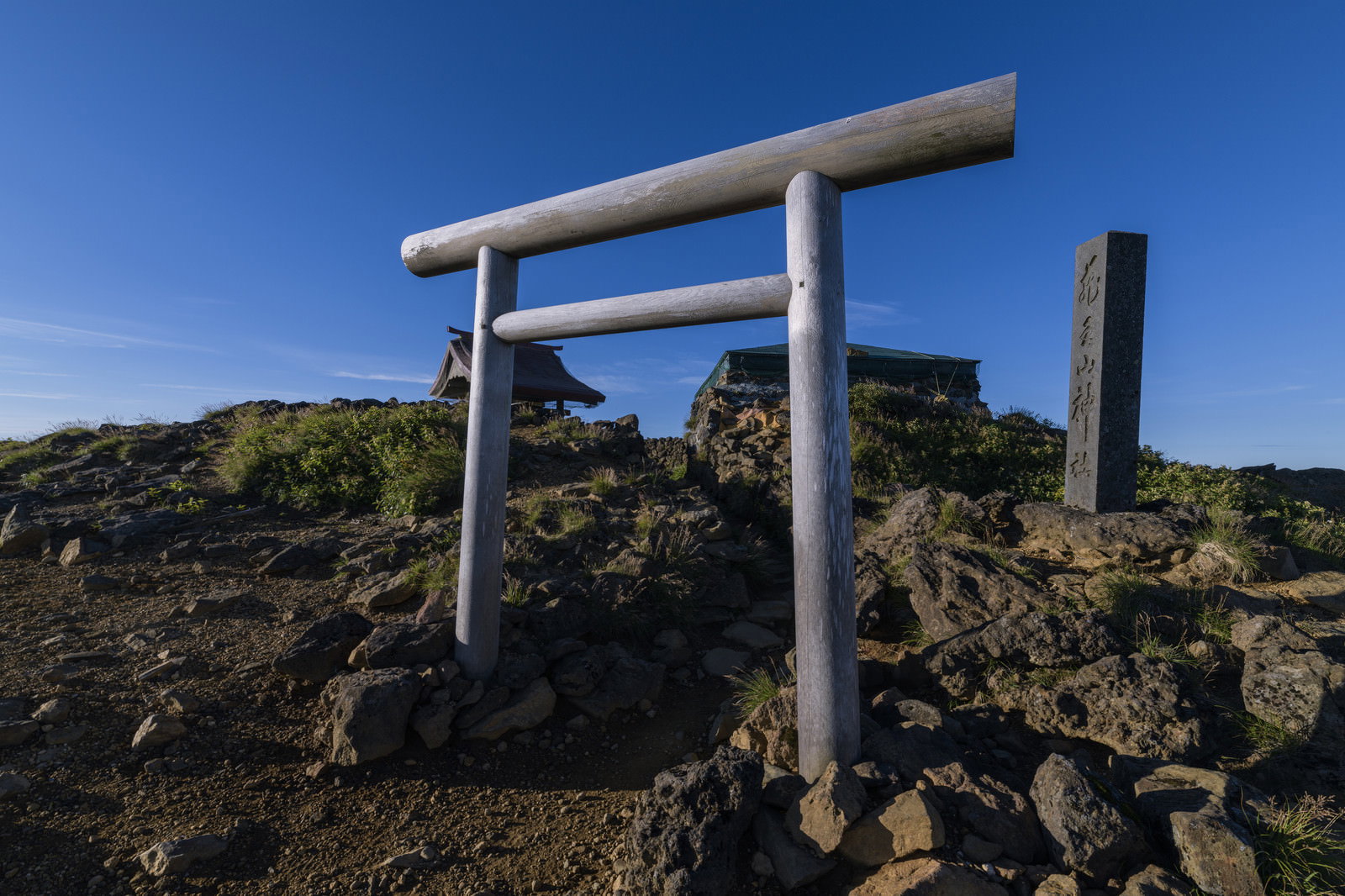 蔵王山の山頂に立つ白い鳥居と青空、背景に石碑が見える山岳風景