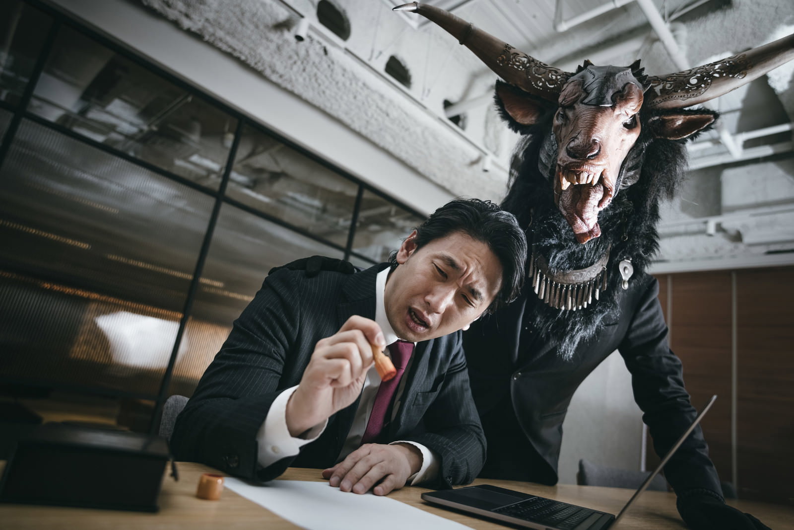 書類を前に困惑するスーツ姿の男性と背後に迫る悪魔の影［モデル：大川竜弥］