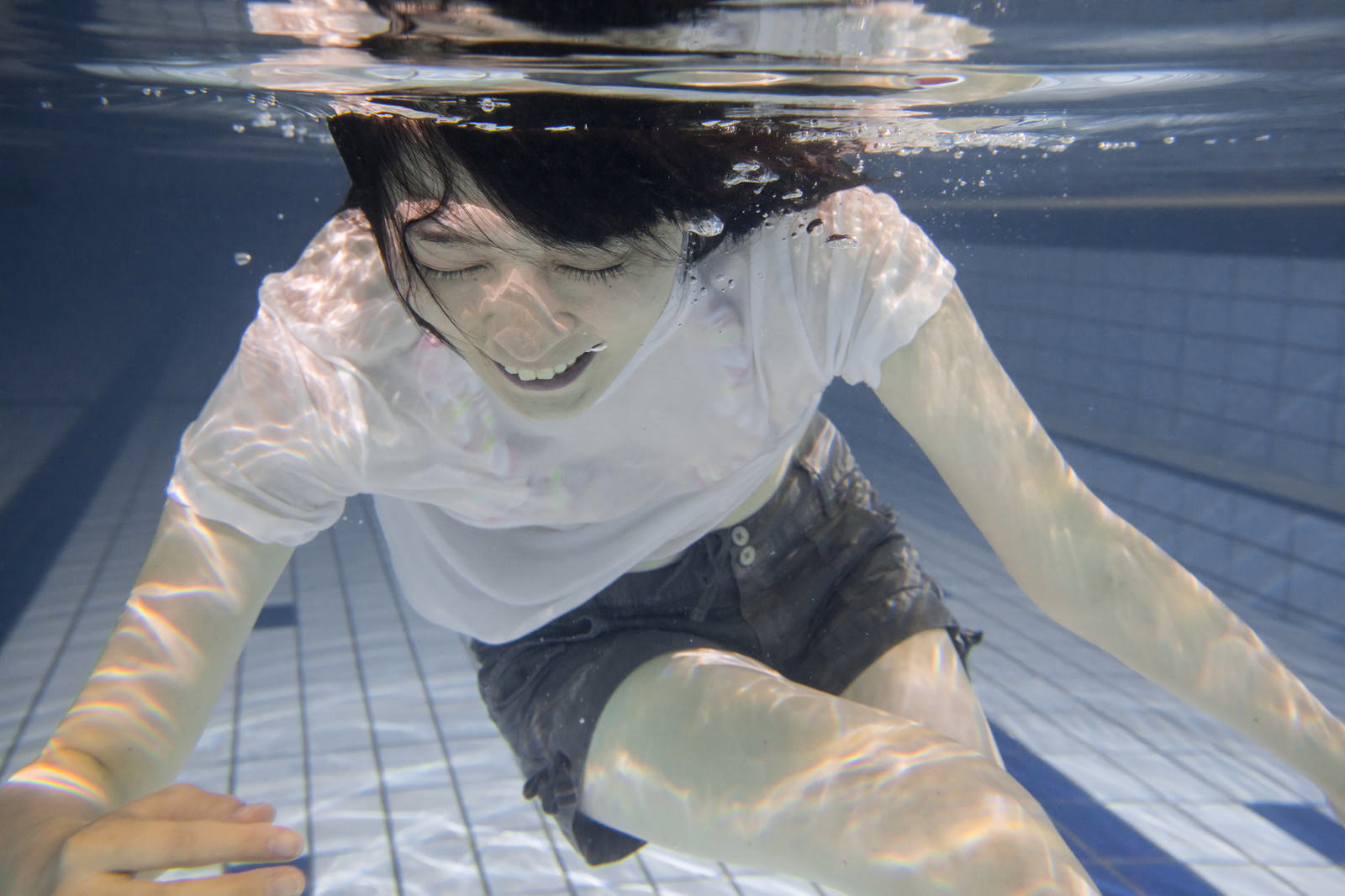 プールの水中で白い水着と帽子を着けた女性が手を上げている［モデル：たけべともこ］