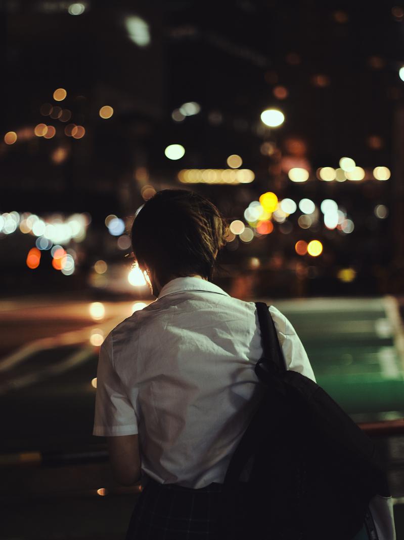 夜の街の灯火を背景に立つ、カバンを背負った帰宅する高校生の後ろ姿