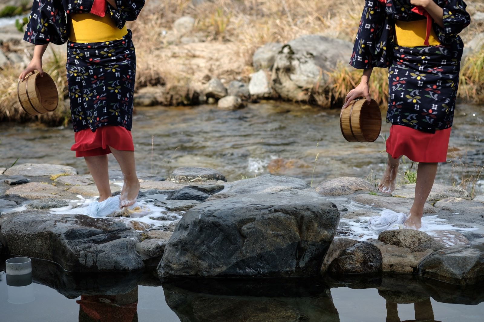 Women in black kimonos with red sashes doing foot-stomping laundry on the stones in the river