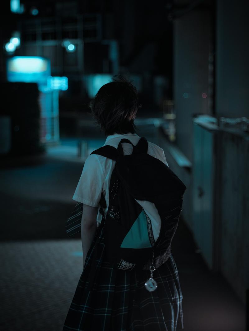 夜の街並みを歩く制服姿の女子高生の後ろ姿。リュック姿で帰路を歩む