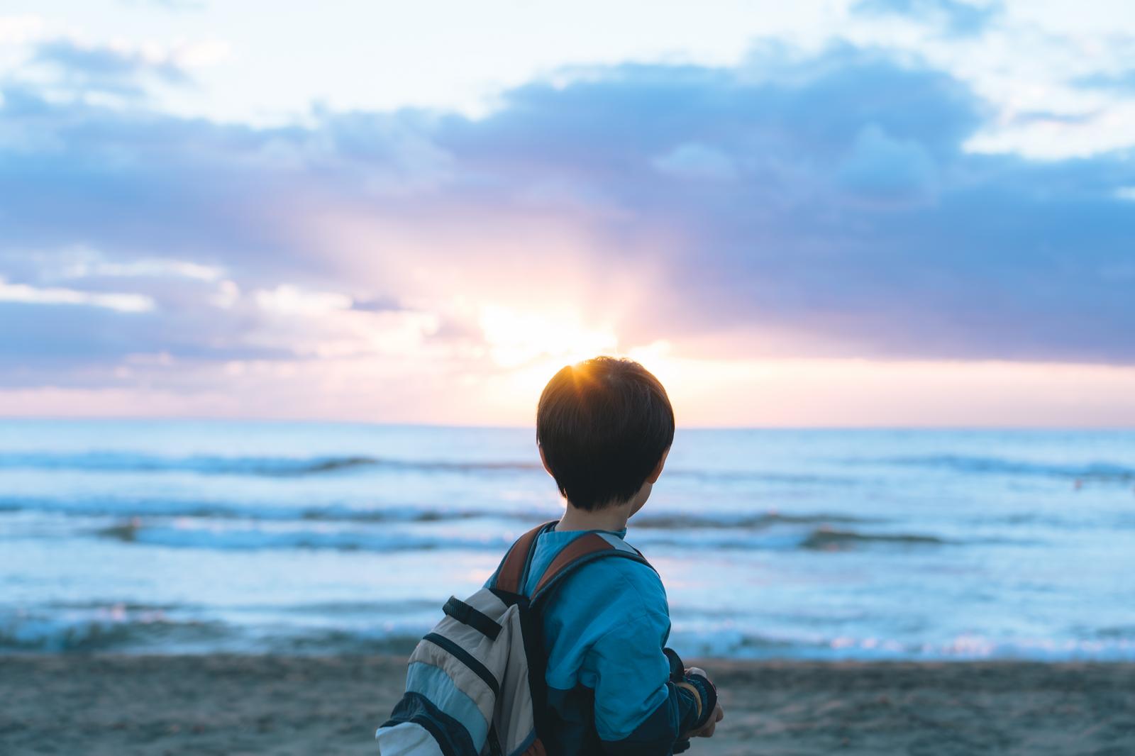 砂浜に立ってリュックを背負い、夕焼けの海を眺める子ども