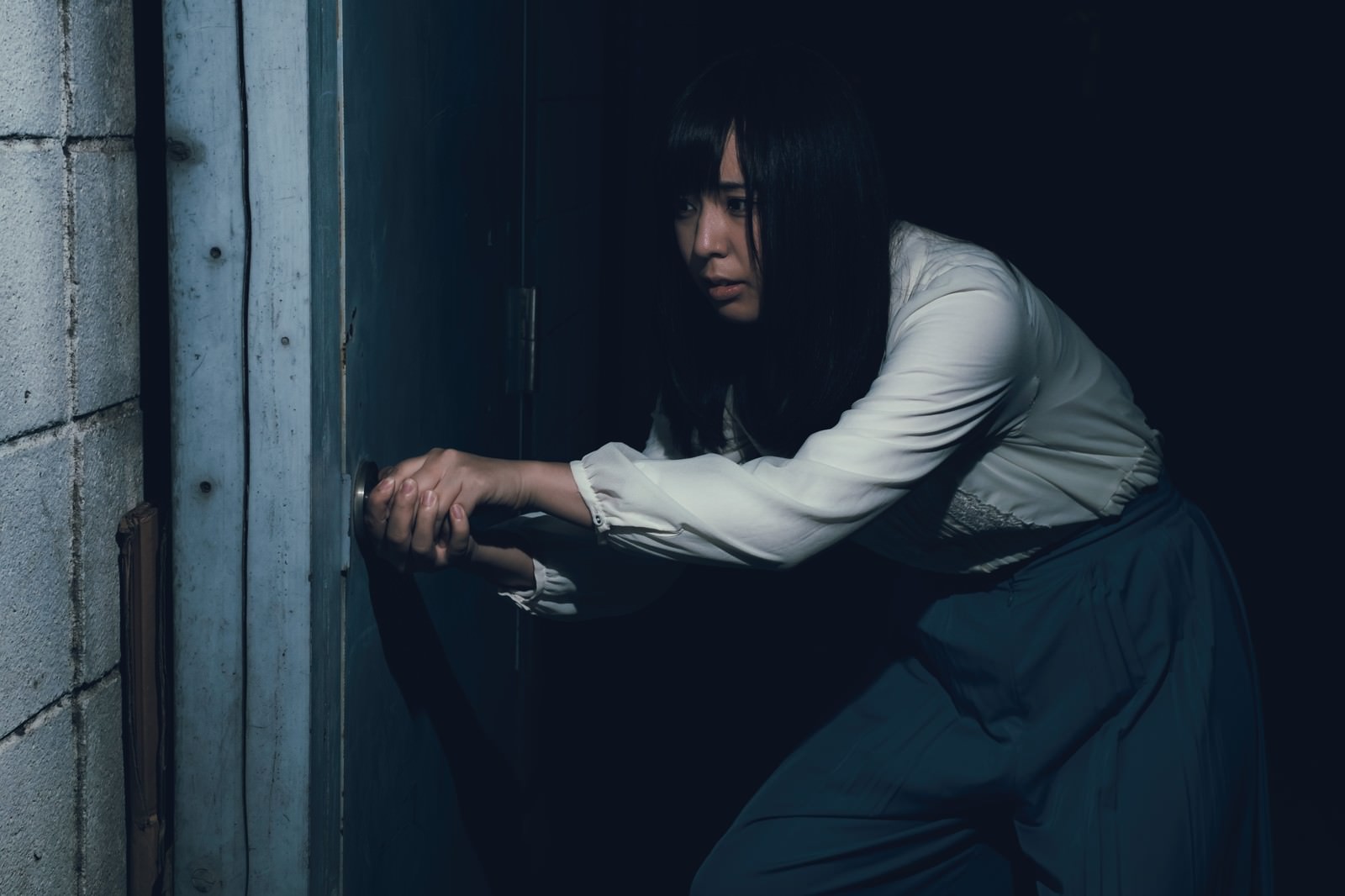 薄暗い室内で古びたドアを恐る恐る開けようとする白い服の女性［モデル：さとうゆい］