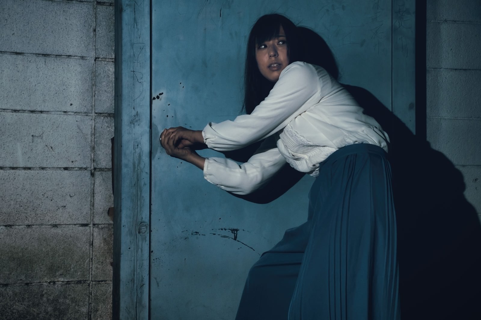 古い青緑色のドア前で白いシャツを着た黒髪の女性がパニック状態で鍵を開けようとしている［モデル：さとうゆい］