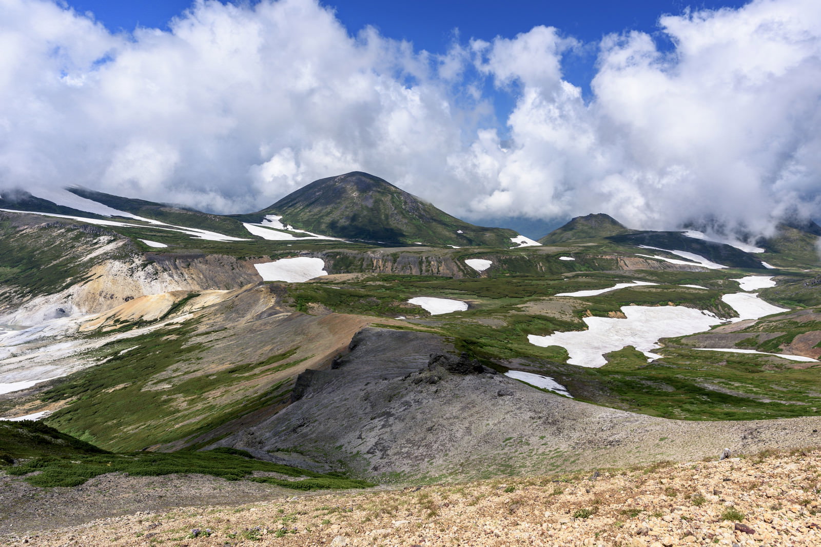 青空と雲の下に広がる大雪山系の山岳風景、残雪と高山植物が点在する