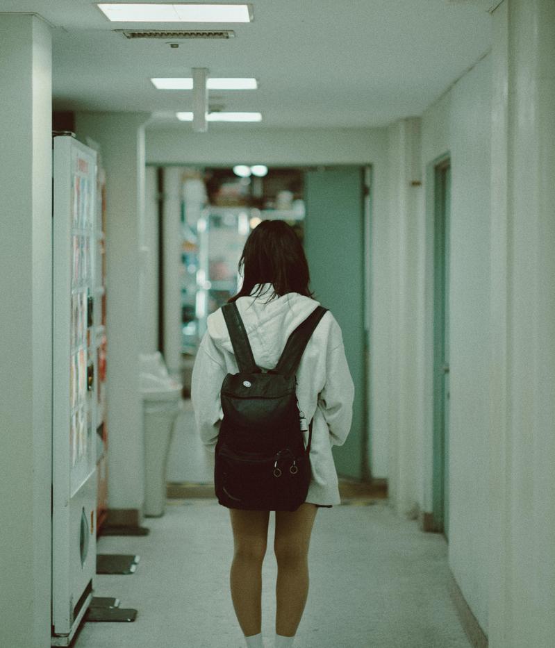 白いシャツに黒いリュックを背負い、廊下を歩く女子高生の後ろ姿
