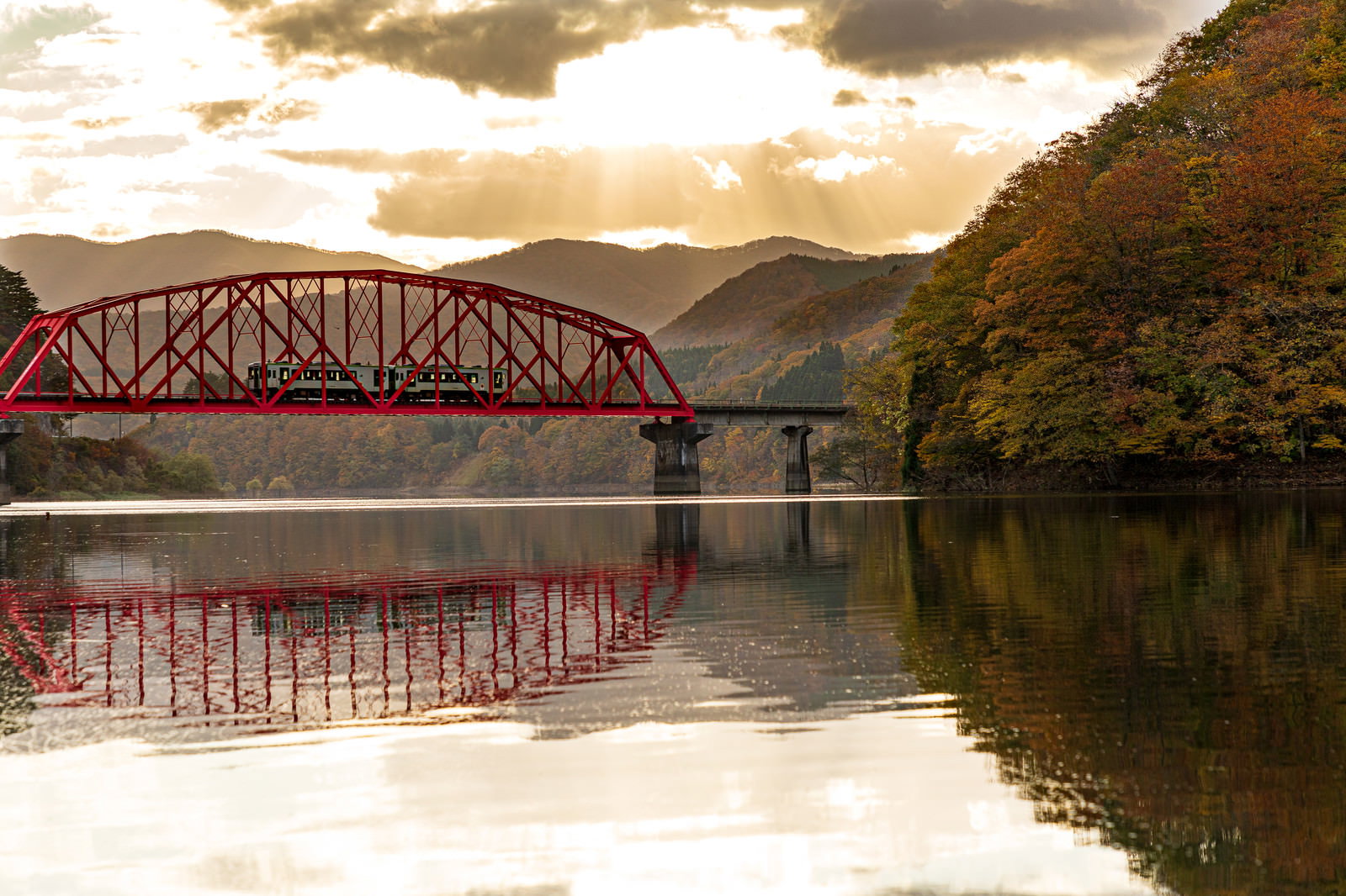 穏やかな湖面に映る赤い鉄橋と電車、周囲に山々と緑の木々