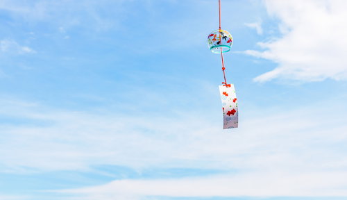赤い紐の風鈴が風で揺れる夏の日中、青空の下で涼しげに音色を響かせる