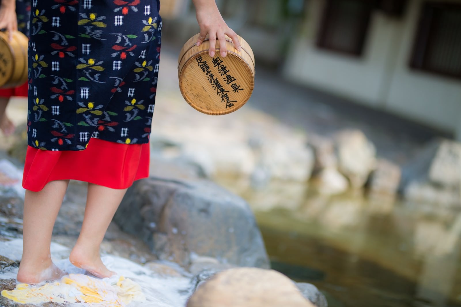 川の中で足踏み洗濯をする紺色花柄ワンピースと赤いスカート姿の女性が木製の洗濯板を手に持つ