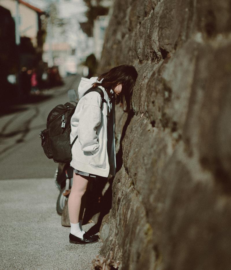 古い石壁に体を預けてうなだれる女子高生、背中に黒いバックパック