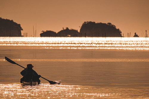 朝日の中を漕ぎ出すカヌーのシルエット、水面がキラキラと輝く